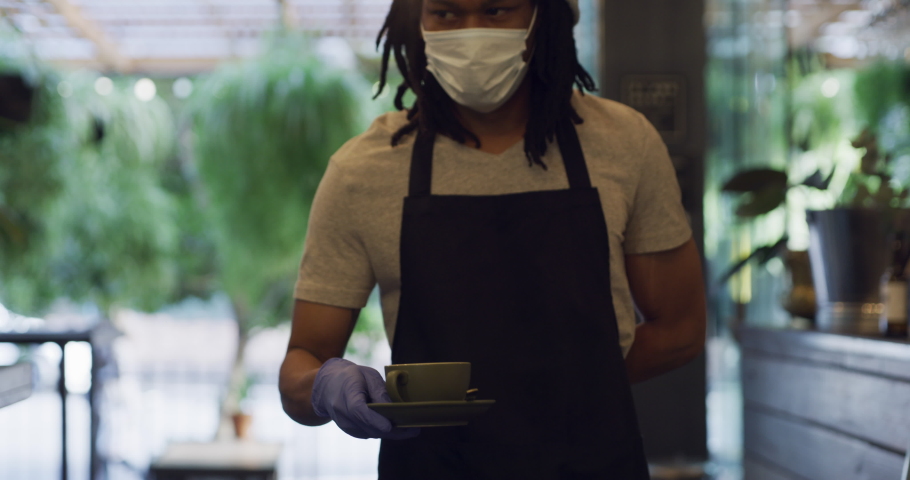 Covid and a waiter is serving a freelance businessman or customer coffee and wearing a covid19 mask at a cafe. A restaurant employee brings tea to a working guy during the covid 19 pandemic Royalty-Free Stock Footage #1097806423