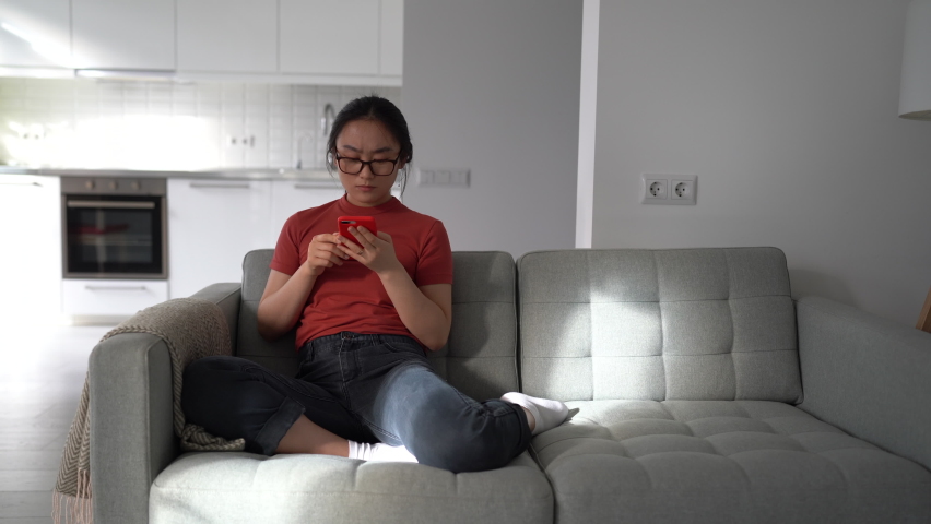 Young unhappy Asian woman looking at mobile phone screen reading bad news throwing away glasses. Upset Korean girl sitting on sofa holding smartphone receiving breakup text message feeling sad | Shutterstock HD Video #1097806901