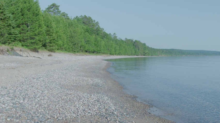 Push in above white pebble beach of forested coastline of Lake Superior, Michigan in summer time