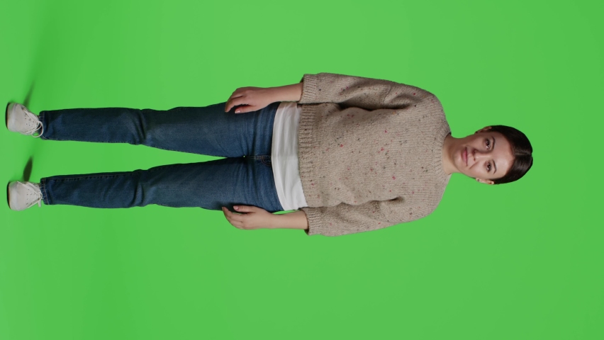 Vertical video: Front view of model standing casual and impatient on camera, posing on greenscreen background. Young woman over isolated green screen backdrop, being thoughtful and waiting, full body.