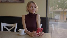 Copy space video of a smiley mature blonde lady using cellphone and drinking a coffee in a cafeteria waiting for her online date. Concept of flirting using mobile app.