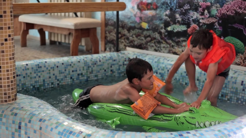 two boys in sleeves and a vest in the pool sit on an inflatable crocodile and laugh