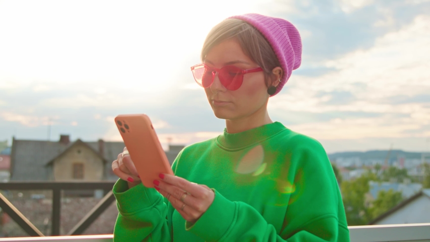 Footage of bright girl in colorful clothes holding smartphone in hands, checking social network. Thoughtful good-looking hipster woman in stylish green sweatshirt looking at screen of device. Oudoors