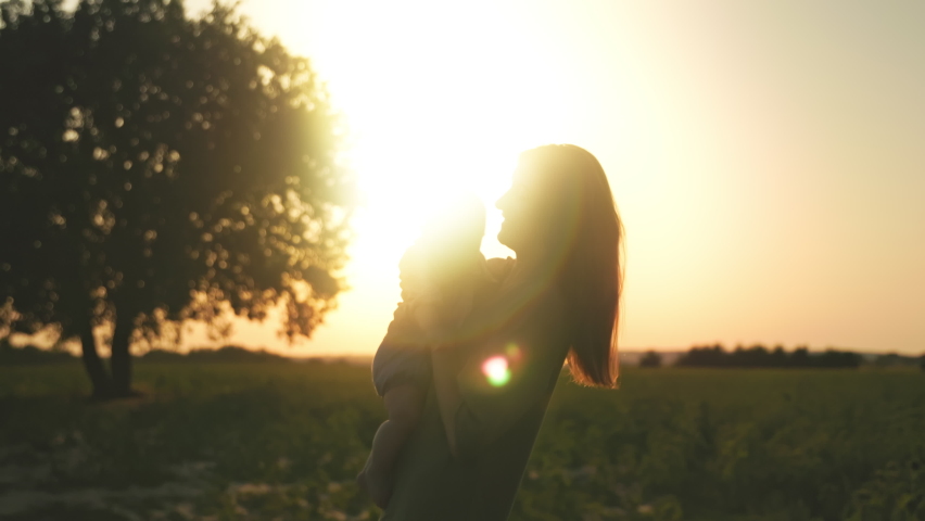 Horizontal view of young happy Caucasian woman holding lovely little baby girl in hands in background of beautiful sunset. Silhouettes of mother and child. Outdoor, field. Village landscape Royalty-Free Stock Footage #1097824991