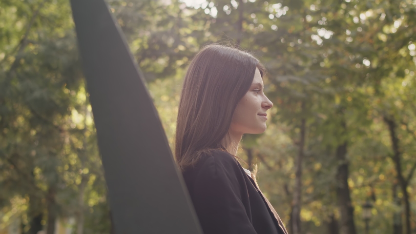 Side view of a beautiful young woman sitting and waiting for her boyfriend outdoors. Man sneaking up on her, putting his fingers over her eyes. Female surprised, laughing
