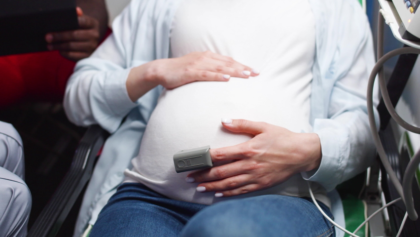 Close-up of pregnant woman pushing during labor suffering from contractions in ambulance with professional African-American and caucasian paramedics listening to heartbeat of baby with stethoscope.