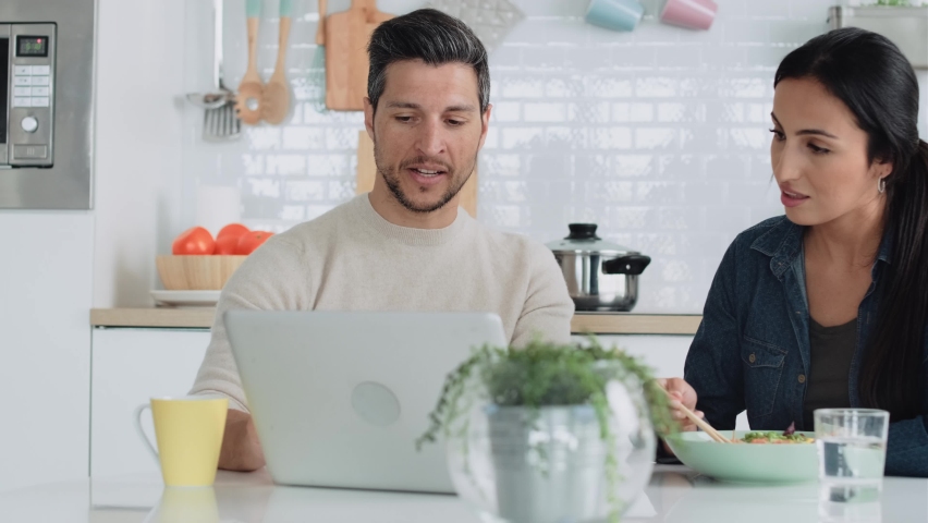 Video of beautiful lovely couple using their laptop while eating noddles with chopsticks in the kitchen at home.
