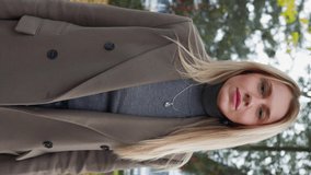 Vertical video, Attractive Business Woman with Blond Hair in Long Coat Walks in Autumn City Park on Cloudy Day. Pretty Lady Blonde Walks and Smiles While Enjoying a Walk in Fall Time. Slow Motion. 