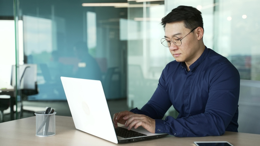 Busy IT specialist programmer with glasses working typing coding on laptop computer in modern office centre Asian entrepreneur professional developer thinking analyzing finance digital data indoors Royalty-Free Stock Footage #1097838281