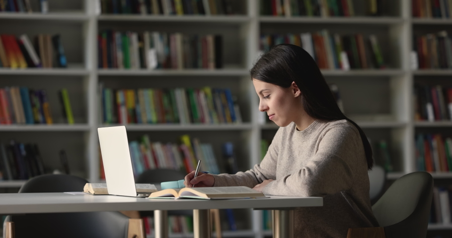 Attractive focused student girl preparing for session, college admission or university exams using laptop, writing information, studying seated at desk in library, side view. Education, modern tech Royalty-Free Stock Footage #1097839769