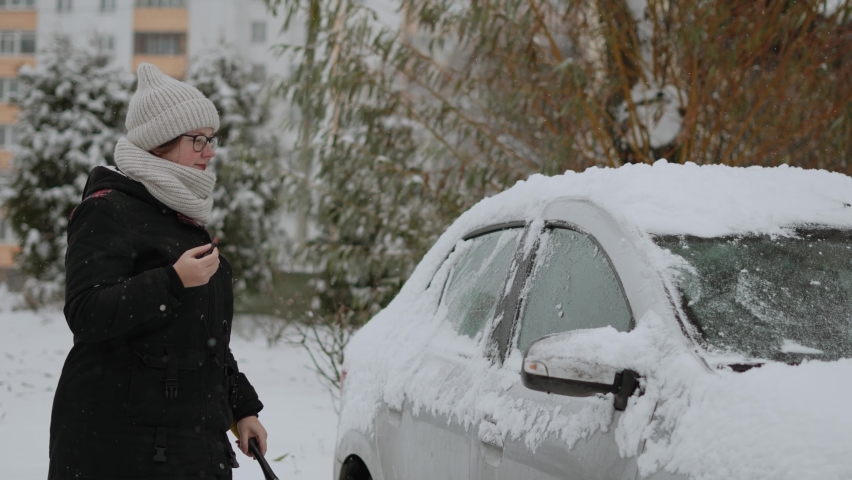 Pretty woman in warm clothes cleans snow from car and paints lips with lipstick. Lady is preparing car for driving in parking lot on frosty winter day. Concept do several things at once..