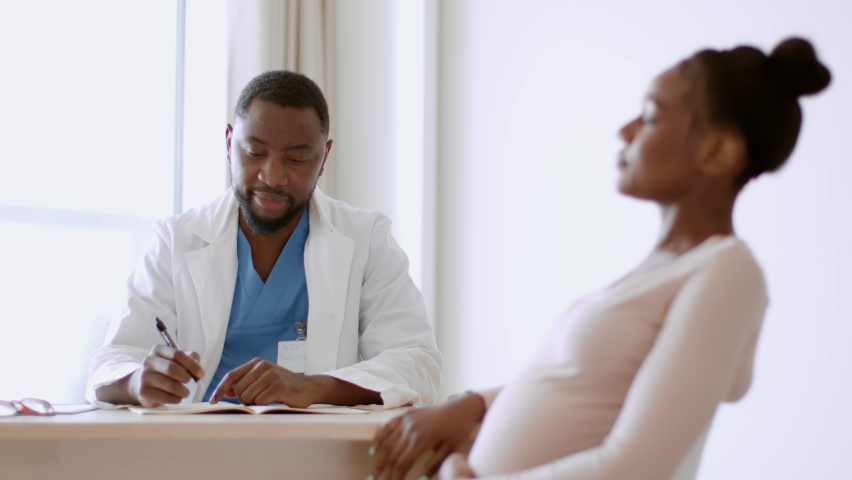 Pregnancy checkup. Young pregnant african american woman sitting at doctor's appointment, professional therapist taking notes in medical history, tracking shot, slow motion, selective focus Royalty-Free Stock Footage #1097844581