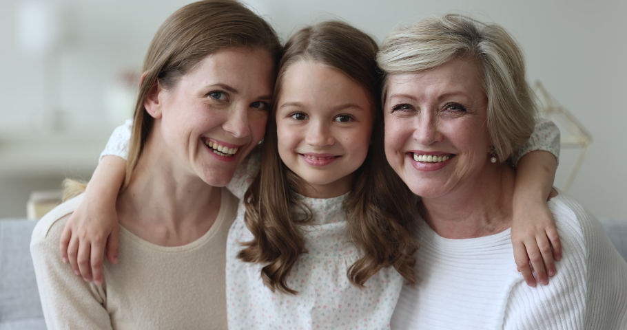 Close up portrait of three generational beautiful women smile look at camera, little girl hugging young mom and older granny sit indoors, feeling unconditional love appreciate each other. Family ties Royalty-Free Stock Footage #1097845035