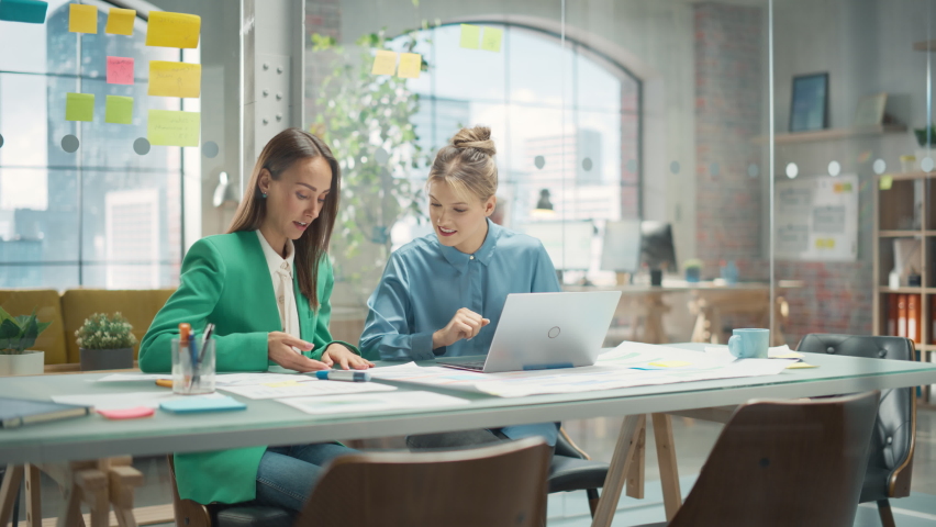 Two Colleagues Working On Laptop in Office. Female Help Desk Coordinator Collaborates with Caucasian Customer Service Agent, They Discuss a Project, Chat, Smile. Teamwork Collaboration