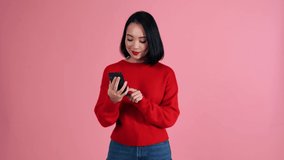 Positive Asian woman wearing red sweater talking by video call on mobile in the pink studio