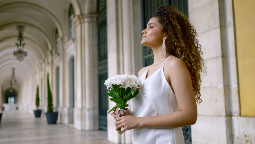 Beautiful black woman in elegant white dress. Action. Attractive woman in white dress with bouquet at old building. Attractive young woman is waiting for boyfriend with flowers