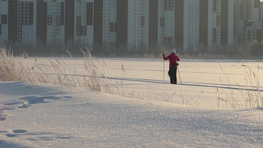 An active woman in a colorful winter jacket is skiing on a large snowy plain along the city houses. freezing day.