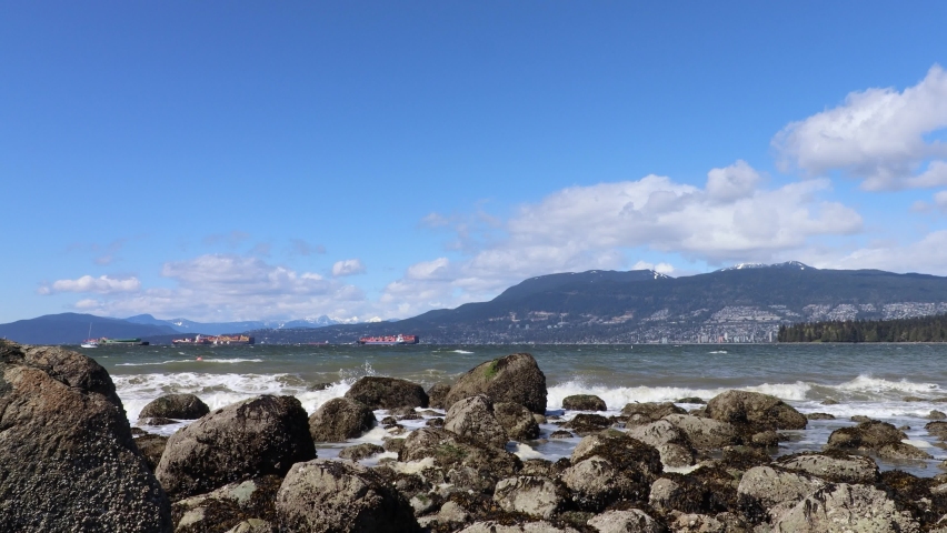 Cargo ships moored far with mountains behind and waves hitting rocks on shore