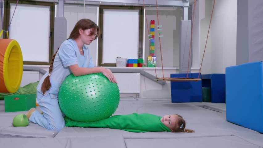 Physical therapist working with little girl in sensory room. Exercising with fitness ball and pressure to help kid relax in a therapy center. sensory integration session Royalty-Free Stock Footage #1097880631