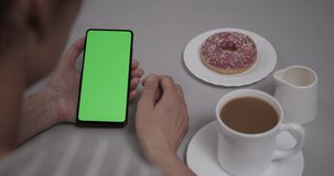 Woman Sitting at Table Coffee Donut Using Smartphone With Chroma Key Green Screen, Scrolling Through Social Network Media Online Shop Internet. Smartphone in Horizontal Mode with Green Screen Mock-up.