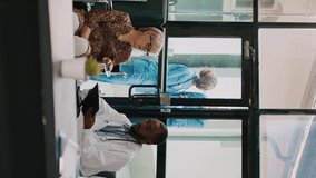 Vertical video: African american doctor consulting old woman in wheelchair, talking about physical disability and disease in hospital waiting room. Specialist and patient with impairment doing medical