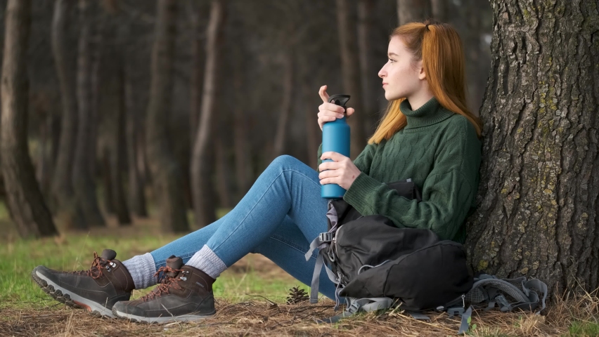 Hiker redhead woman pulling a water bottle from backpack and drinking sitting under tree in forest. Trekking concept.