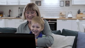 Modern cheerful caucasian mother grandmother holds adorable little son in arms. Uses laptop computer in living room kitchen in apartment. Watches funny video on social networks and apps on screen