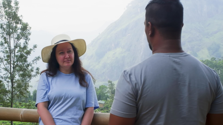 Woman and man are talking in nature park with mountains standing on bridge. Woman is looking at him, smiling and listening, He is standing back to camera. Tourism, travel, trip, vacation together.