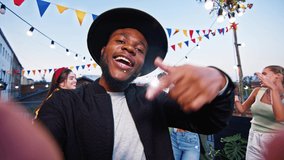 POV of young handsome happy African-American man looking at camera singing on rooftop having party with young people having fun dancing. Attractive friends spending time on music event.