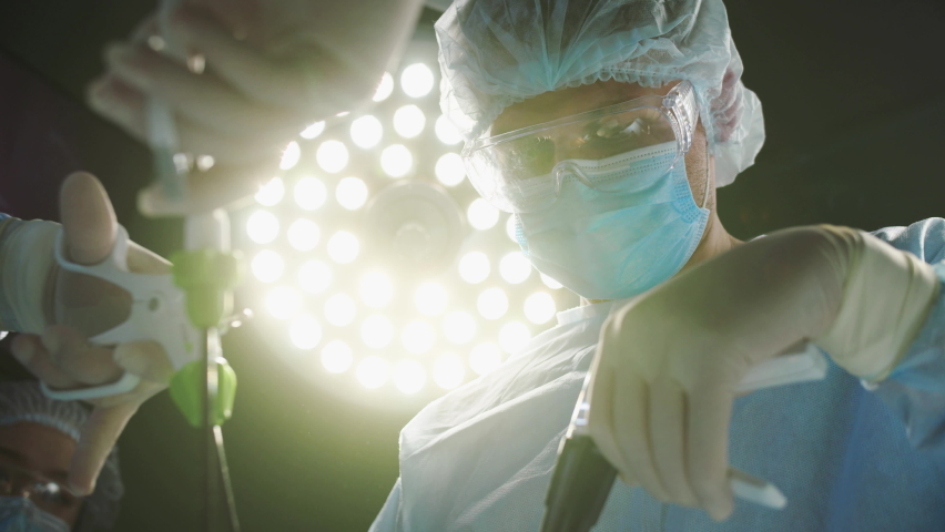 POV low angle view of patient being operated by qualified surgeons wearing medical gowns, caps, masks and gloves. Group of professional doctors using modern medical equipment in operation room. Royalty-Free Stock Footage #1097905453