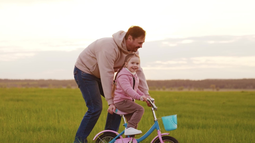 happy family. father teaches child ride bike sunset. cheerful kid controls steering wheel bicycle ray sunset light sun. daughter kid child rides next dad green lawn. child dream kid. childhood parent. Royalty-Free Stock Footage #1097906233
