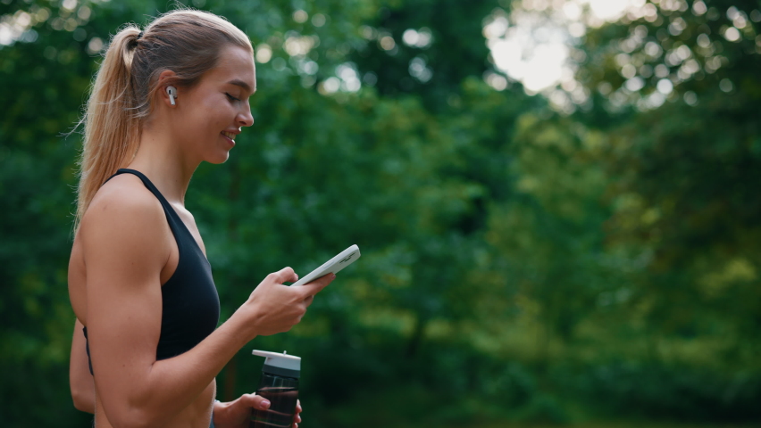 Side view of the sporty woman walking, holding sport bottle using, scrolling phone and listening to music using earphones. Sport and technology and healthy lifestyle concept Royalty-Free Stock Footage #1097906789