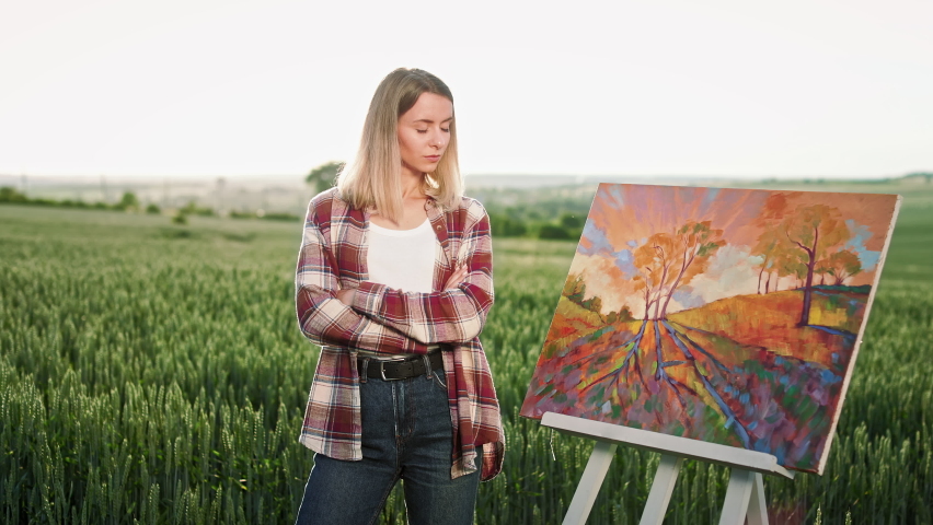 Front view of pretty, blonde woman showing picture of autumn landscape. Cheerful woman wearing plaid shirt and jeans, painting, holding brush and palette of colors. Concept of art.