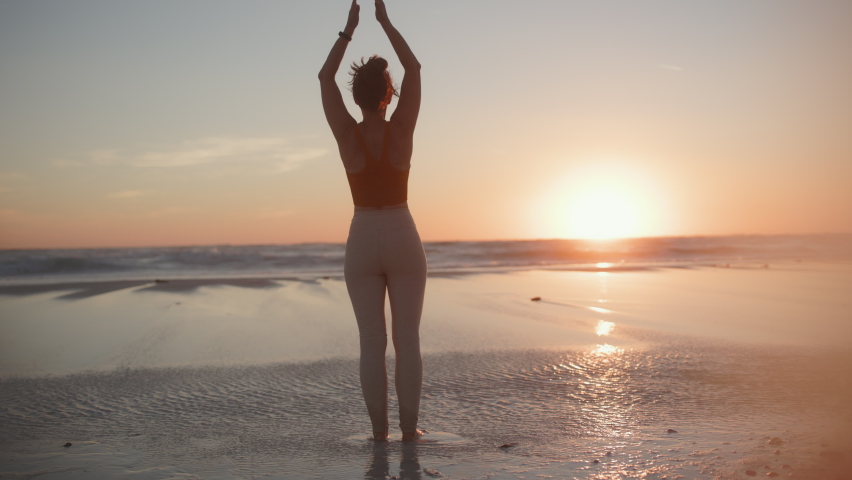 Seen from behind fitness woman jogger in fitness clothes at the beach at sunset meditating.
