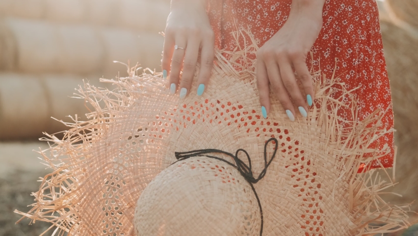 Woman in red dress holding in hands straw trendy hat. Girl with a straw hat in her hands stands against the background of haystacks. Concept: Happiness, Travel, Lifestyle, Freedom. 