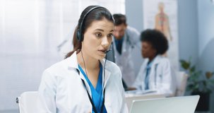 Close up of Caucasian young beautiful female doctor having online video call consultation on laptop speaking in headset with patient sitting at workplace in hospital cabinet. Healthcare, online chat