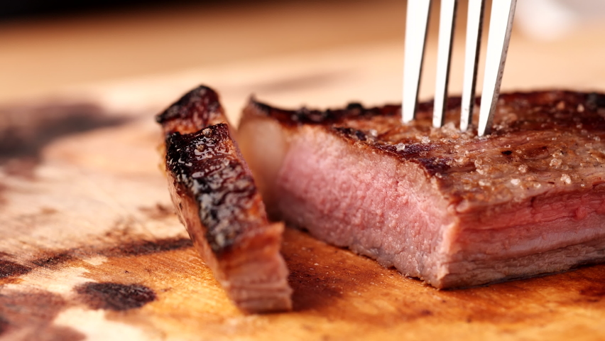 Chef slicing medium or well done beef steak with knife and fork on wooden cutting or chopping board, 4K. Concept of meat, beef steak