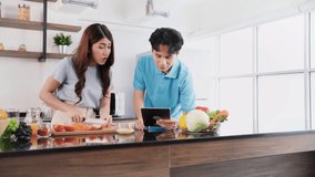 Happy Asian couple cooking healthy food. Man using a digital tablet to search and check the recipe during woman prepare a vegetarian healthy salad with fresh vegetables in the home kitchen.