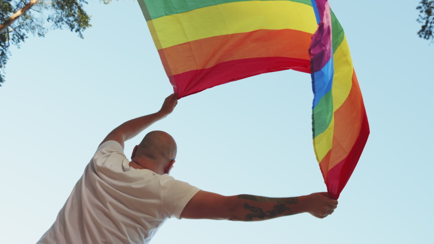 Queer man holding rainbow gay flag while parade on background of blue sky. Happy guy wearing heart sunglasses demonstrate his rights. LGBTQI, Pride Event, LGBT Pride Month, Gay Pride Symbol Royalty-Free Stock Footage #1097949659