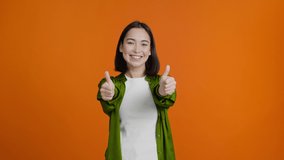 Positive young woman shows thumbs-up. Happy Asian female in green shirt smiles cheerfully. Glad brunette lady expresses approval against orange wall - Powered by Shutterstock - Get 15% off with code: PIKWIZARD15