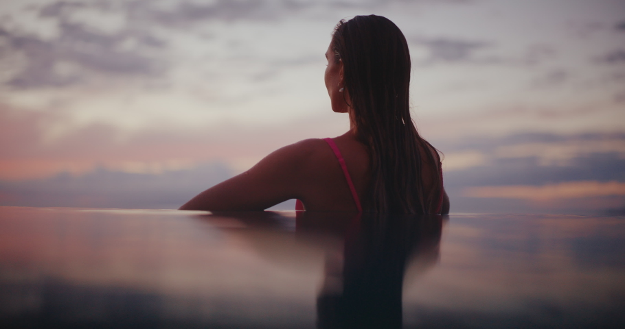 Beautiful young woman relaxing on the edge of infinity pool at luxury hotel resort spa, looking out over the ocean at sunset on vacation Royalty-Free Stock Footage #1097954761