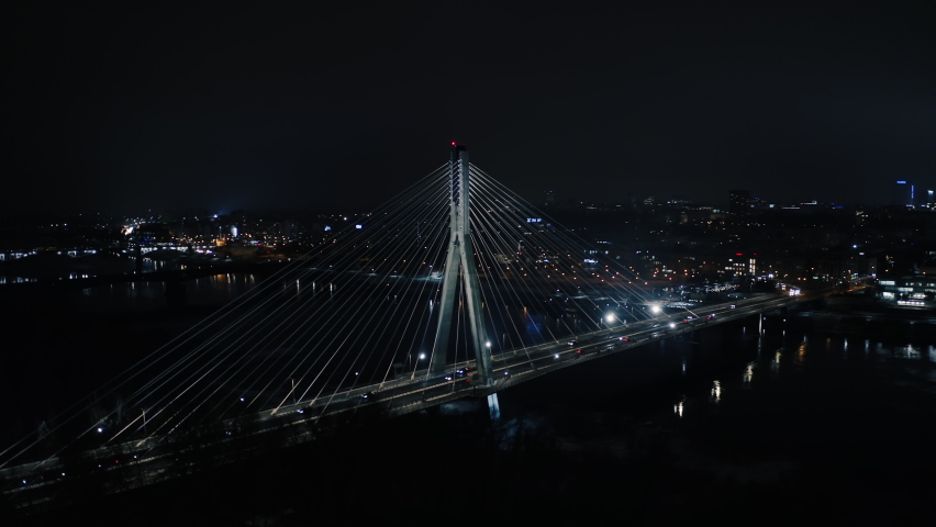 Aerial panoramic shot of Warsaw cityscape. Swietokrzyski suspension bridge over Vistula river, skyscrapers downtown urban skyline at night. Cinematic city panorama flyover cable bridge towers