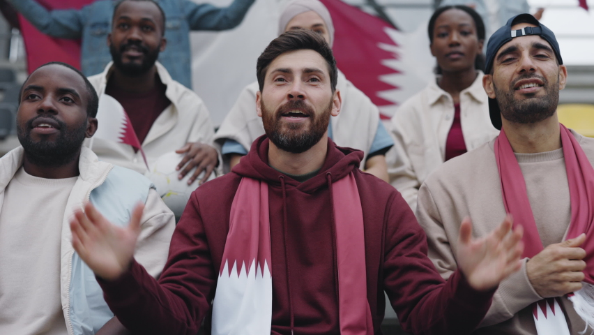 Bearded caucasian man gesturing and screaming while cheering favorite team during soccer game. Devoted fan sitting on tribune with another multiracial spectators.