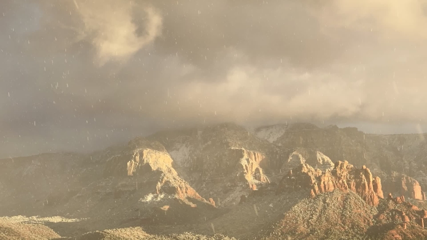 Multiple red rock peaks with snow and low clouds and sun and snow blowing in to a near white out
