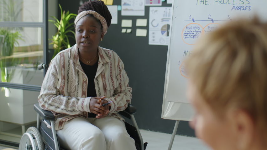 African American businesswoman in wheelchair explaining scheme on flipchart while giving presentation to colleagues during office meeting | Shutterstock HD Video #1097965429