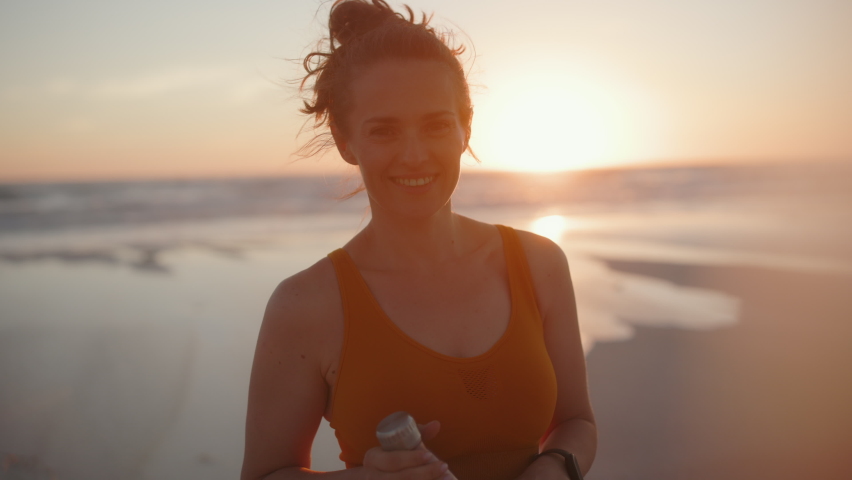 smiling fitness woman jogger in fitness clothes with bottle of water at the beach in the evening.