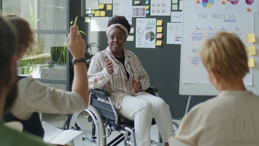 African American female business coach with disability sitting in wheelchair and answering questions about scheme on flipchart while teaching employees in office | Shutterstock HD Video #1097968991