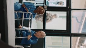 Vertical video: Female nurse doing checkup consultation with old patient in hospital waiting area, having walking frame. Medical assistant writing clinical report or appointment form, health center.