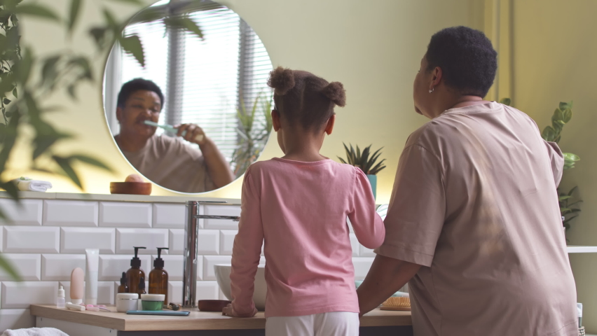 Back view of African American woman and her little daughter standing by sink in front of mirror in bathroom brushing their teeth