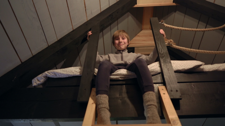 Happy little boy is sitting on a bed on a second floor under a roof in a little cozzy wooden house. Winter vacations concept
